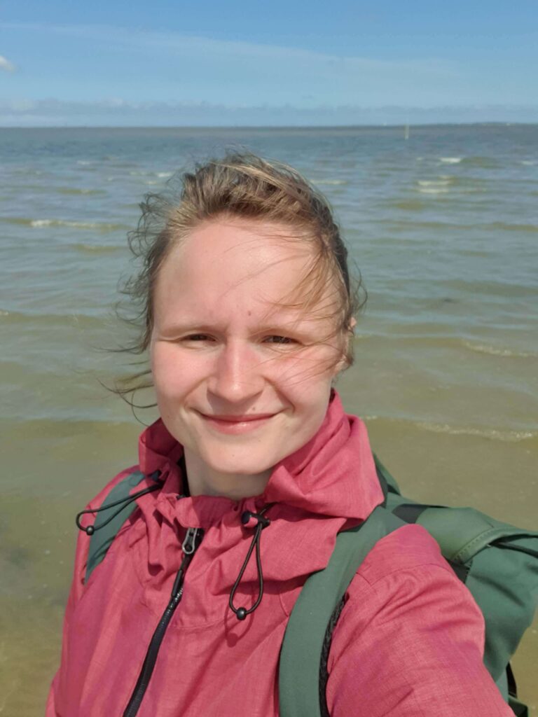 Jacqueline unterwegs am Meer- Reisen mit Camper und Hund in Europa. Es ist windig, die Haare sind verweht aber die Sonne scheint. Im Hintergrund ist das Meer zu sehen.