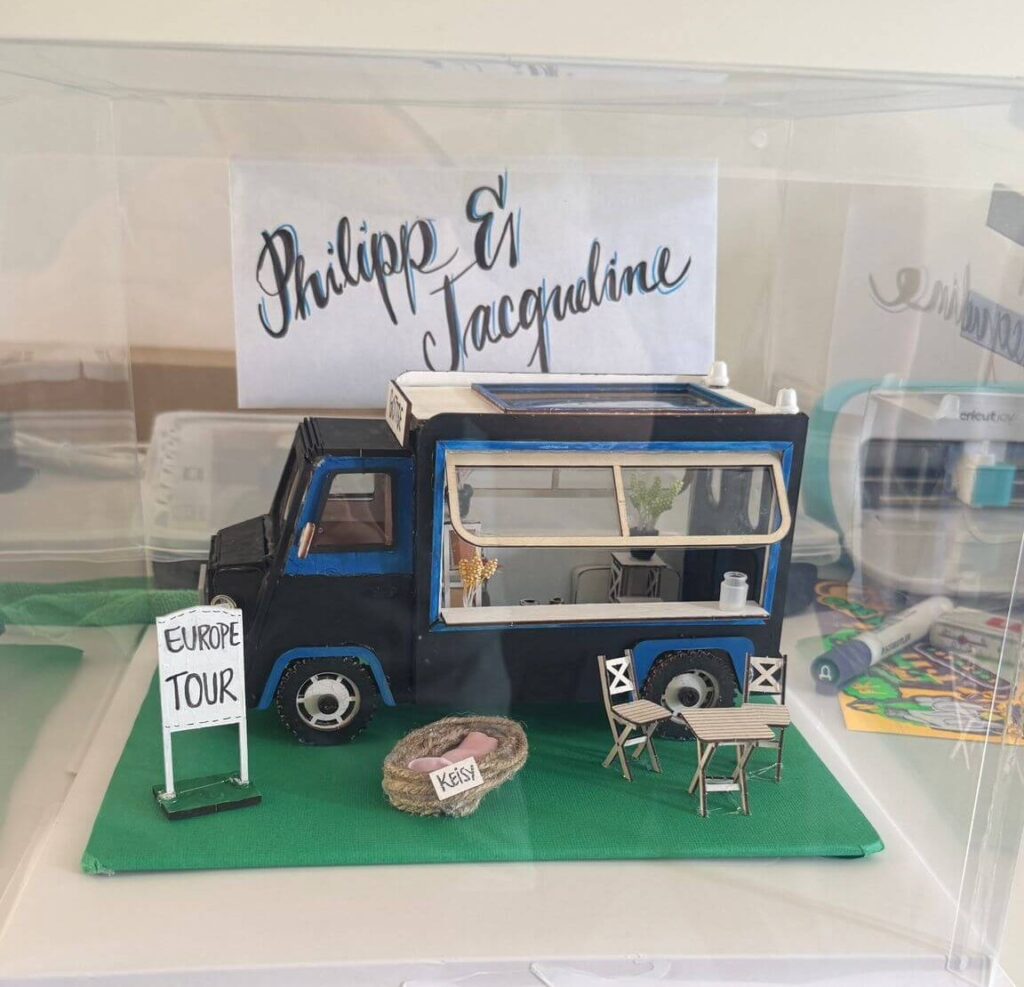 Ein liebevoll gestaltetes Miniatur Model eines schwarz blauen Campers mit einem Schild Europe Tour und einem Hintergrund Schild mit den Namen Philipp& Jacqueline. Unser besonderes Hochzeitsgeschenk.