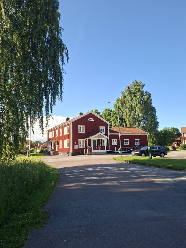 Großes rotes Holzhaus mit weißen Fenstern in einer schwedischen Ortschaft. Es scheint die Sonne und der Himmel ist Wolkenlos. Abschnitt über den Holz kauf in Schweden sowie häufige Fragen zum Immobilienkauf.